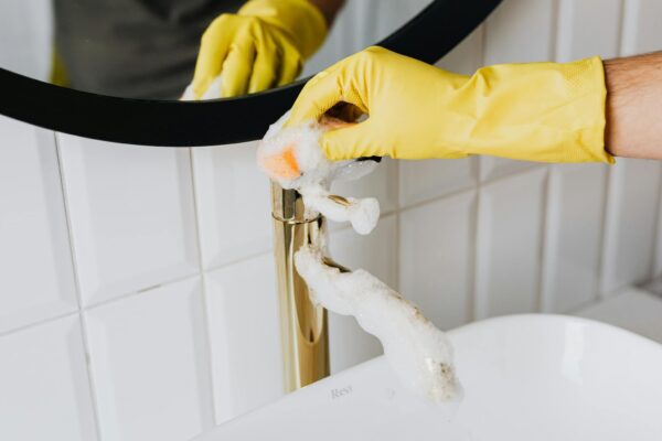 Anonymous person wearing yellow latex gloves washing golden faucet with sponge in modern bathroom while cleaning house