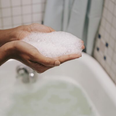 Crop anonymous female with bubbling foam in hands over bathtub while washing in bathroom