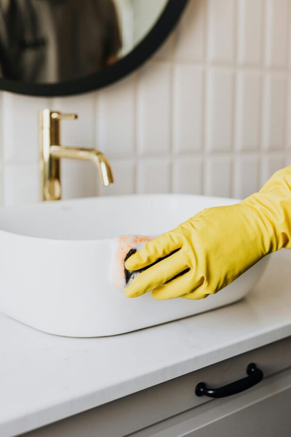 Crop faceless person in yellow rubber protective glove wiping surface of white sink in bathroom with sponge and cleaning agent